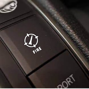 May include: A black car dashboard with a white button labeled "FIRE" and a compass icon. The button is located next to a button labeled "SPORT".