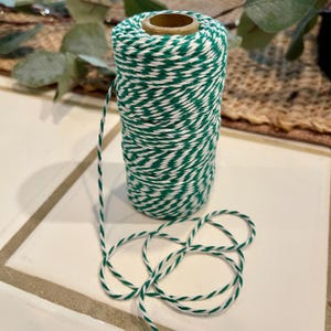 May include: A spool of green and white twine. The twine is tightly wound around a cardboard core. A length of the twine extends from the spool and is looped on the surface. The twine has a twisted pattern of green and white.
