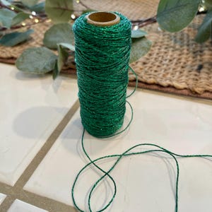 May include: A spool of emerald green twine with a subtle glitter effect. The twine is tightly wound around a cardboard core. The background includes eucalyptus leaves and a woven placemat, suggesting a craft or decorative theme.