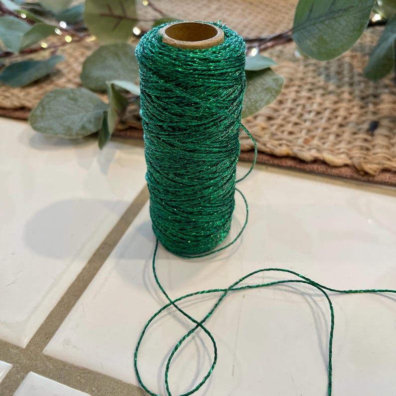 May include: A spool of emerald green twine with a subtle glitter effect. The twine is tightly wound around a cardboard core. The background includes eucalyptus leaves and a woven placemat, suggesting a craft or decorative theme.