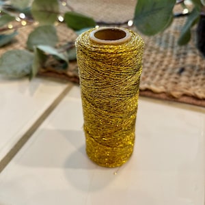 May include: A spool of gold metallic thread. The thread is tightly wound around a cardboard core, creating a cylindrical shape. The metallic thread has a shiny, glittery appearance, reflecting light. This thread is suitable for various crafting projects.