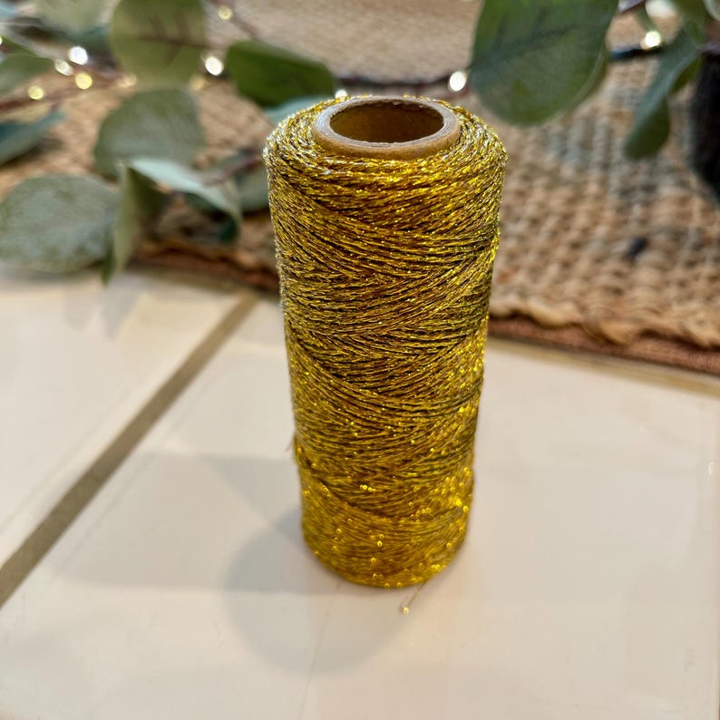 May include: A spool of gold metallic thread. The thread is tightly wound around a cardboard core, creating a cylindrical shape. The metallic thread has a shiny, glittery appearance, reflecting light. This thread is suitable for various crafting projects.