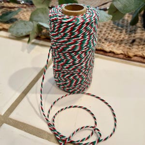 May include: A spool of baker's twine with a festive red, green, and white twisted pattern. The twine is wrapped around a cardboard core. A length of the twine is unwound and lies on the surface.