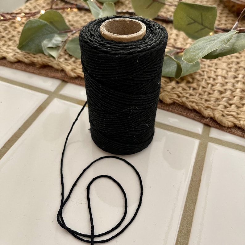 May include: A spool of black twine with a cardboard core. The twine is tightly wound and a length of the twine is unwound, creating a loop. The spool is on a white tiled surface.