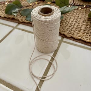 May include: A spool of natural-colored cotton twine. The twine is tightly wound around a cardboard core, with a length of the twine unwound and lying in a loose coil. The twine is suitable for crafts, gift wrapping, or general use.
