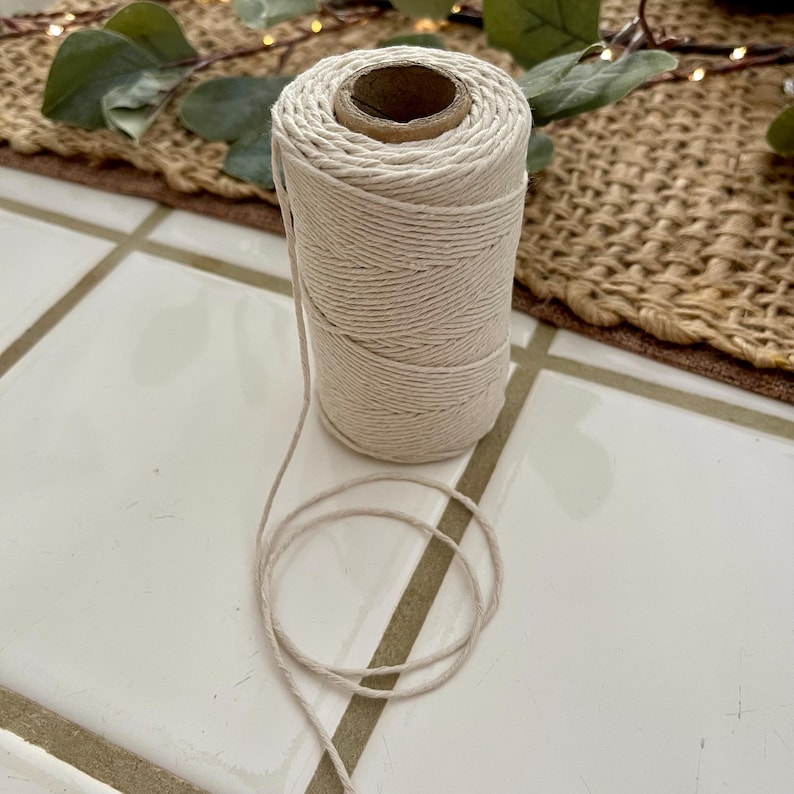 May include: A spool of natural-colored cotton twine. The twine is tightly wound around a cardboard core, with a length of the twine unwound and lying in a loose coil. The twine is suitable for crafts, gift wrapping, or general use.