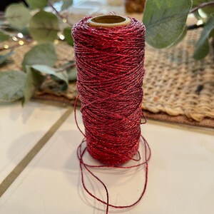 May include: A spool of red glitter twine. The twine is tightly wound around a cardboard core. The red color has a metallic sheen, catching the light. Strands of the twine are loose at the base of the spool. The background includes greenery and a woven surface.