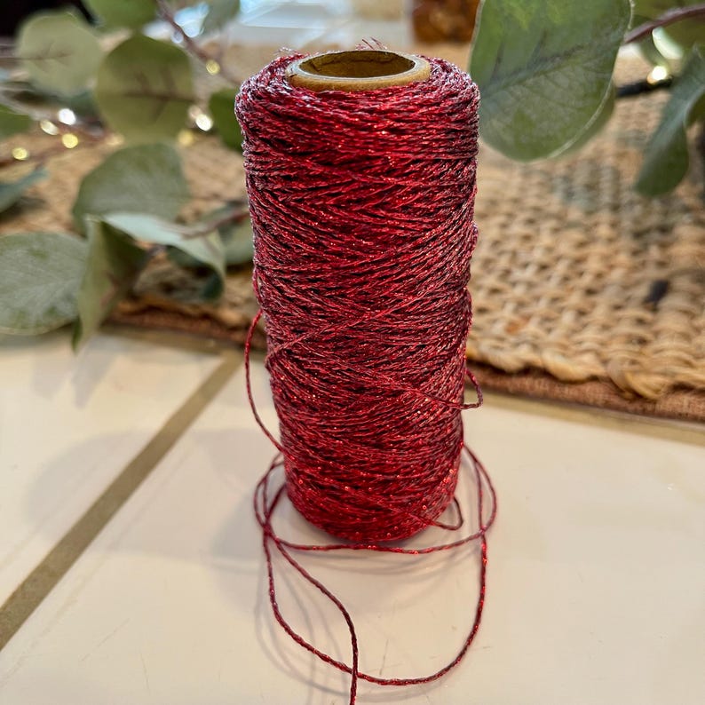 May include: A spool of red glitter twine. The twine is tightly wound around a cardboard core. The red color has a metallic sheen, catching the light. Strands of the twine are loose at the base of the spool. The background includes greenery and a woven surface.