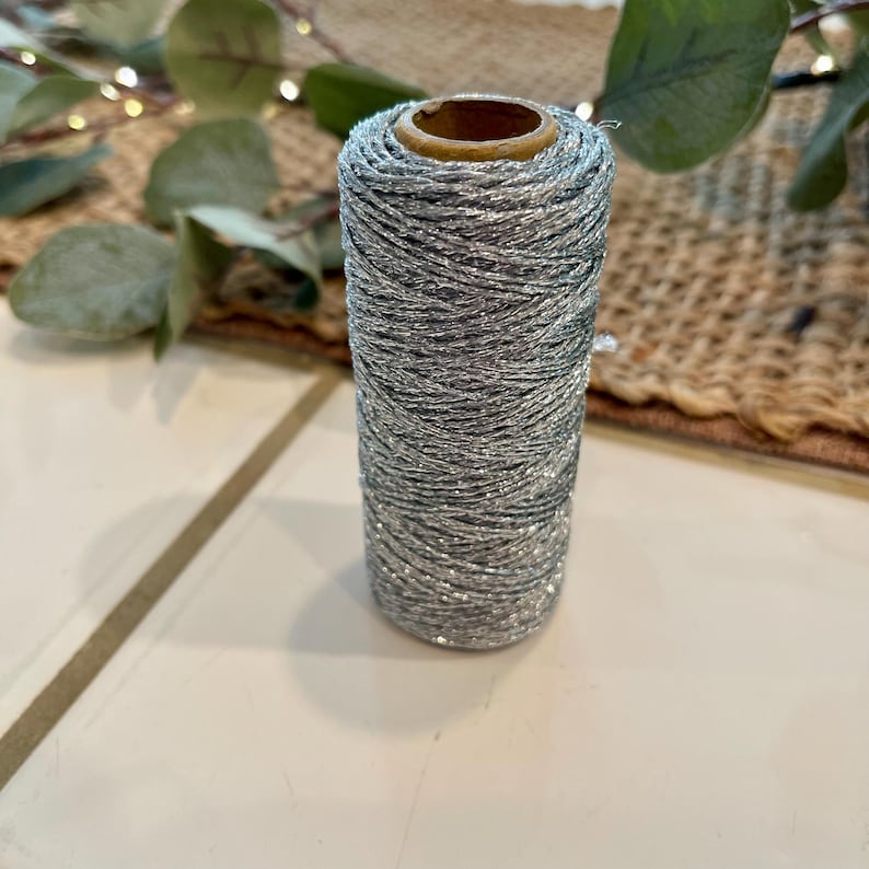 May include: A spool of silver metallic thread. The thread is tightly wound around a cardboard core. The thread has a sparkly, glittery appearance. The spool is standing upright on a white surface.