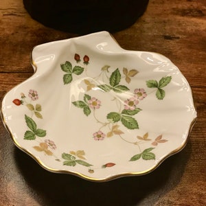 May include: A white porcelain shell-shaped dish with a gold rim. The dish is decorated with a floral pattern of green leaves and pink flowers, and red strawberries.