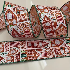 May include: A wired-edge ribbon featuring a repeating pattern of gingerbread houses on a brown background. The houses are decorated with white icing details, red doors, and green accents. The ribbon has a dark green stitched border.
