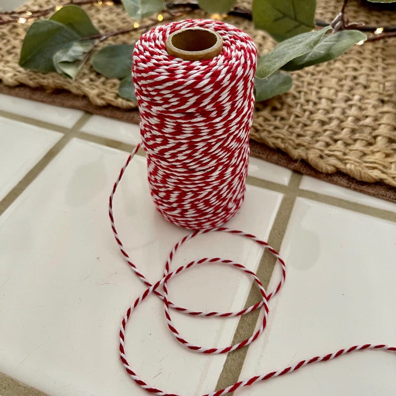 May include: A spool of red and white baker's twine. The twine is tightly wound around a cardboard core. A length of the same twine is unwound and lies in a loop on a white tile surface. Greenery is visible in the background.