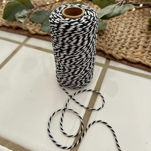 May include: A spool of black and white baker's twine. The twine is tightly wound around a cardboard core. A length of the twine is unwound, forming loose loops on a white tile surface. The twine is suitable for crafts, gift wrapping, and other decorative uses.