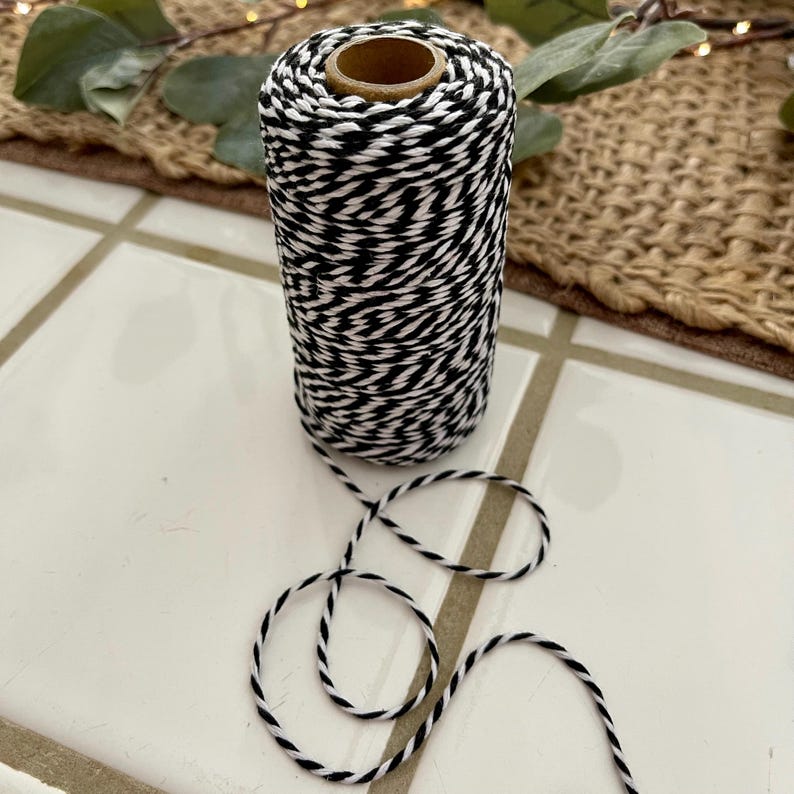 May include: A spool of black and white baker's twine. The twine is tightly wound around a cardboard core. A length of the twine is unwound, forming loose loops on a white tile surface. The twine is suitable for crafts, gift wrapping, and other decorative uses.