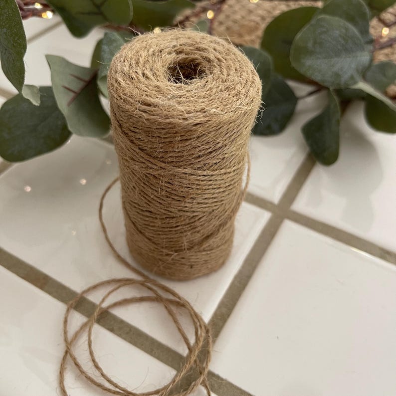 May include: A spool of natural-colored twine sits on a white tile surface. The twine is tightly wound into a cylindrical shape, with loose strands at the bottom. Green leaves and a string of lights are in the background.