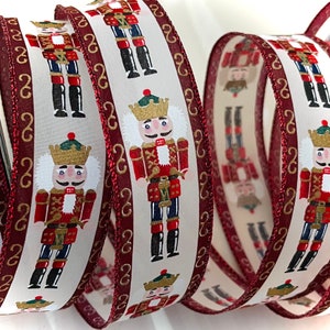 May include: White satin ribbon with a repeating pattern of nutcracker figures in red, gold, and blue. The ribbon has a burgundy edge with gold trim.