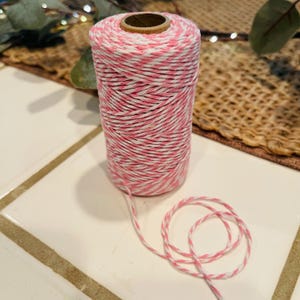 May include: A spool of pink and white baker's twine. The twine is tightly wound around a cardboard core. A length of the twine is unwound and coiled on the surface. The twine is suitable for crafts, gift wrapping, and other decorative uses.