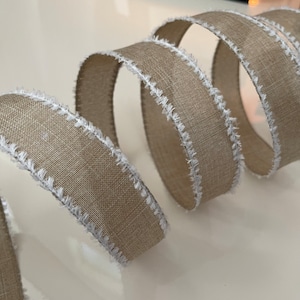 May include: Close-up of a decorative ribbon with a natural tan color and white frayed edges. The ribbon is curled, showing its texture and design. Ideal for crafts and gift wrapping.