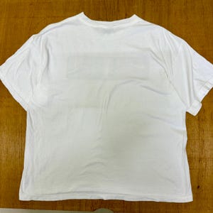 Op de afbeelding: Een wit T-shirt met korte mouwen. Het shirt is gemaakt van een zacht materiaal en heeft een ronde hals. De achterkant van het shirt heeft een rechthoekig ontwerp. Het shirt ligt plat op een houten oppervlak.