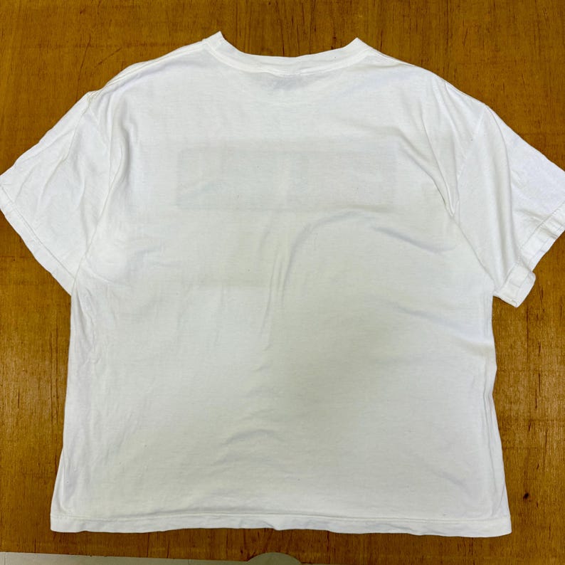 Op de afbeelding: Een wit T-shirt met korte mouwen. Het shirt is gemaakt van een zacht materiaal en heeft een ronde hals. De achterkant van het shirt heeft een rechthoekig ontwerp. Het shirt ligt plat op een houten oppervlak.