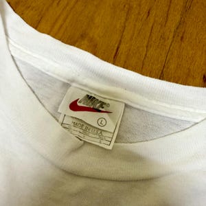 Op de afbeelding: Wit T-shirt met een Nike logo en label. Het label luidt "MADE IN U.S.A." en bevat maat "L". Het Nike logo is rood met een zwarte swoosh. Het shirt is gemaakt van zacht, wit materiaal.