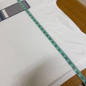 Op de afbeelding: Wit t-shirt met een rechthoekig ontwerp in grijs, wit en blauw. Een meetlint ligt over het shirt en toont metingen in centimeters. Het shirt is gemaakt van een zacht materiaal, waarschijnlijk katoen.