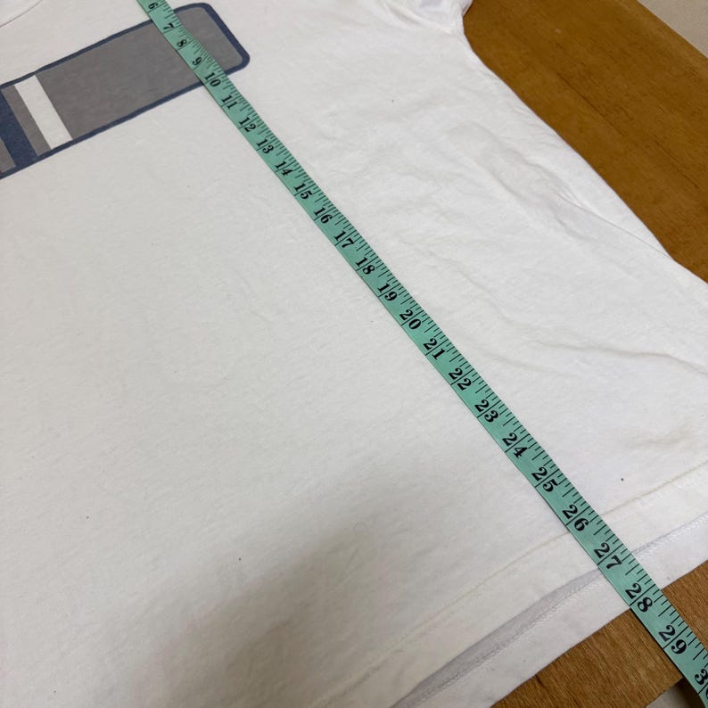 Op de afbeelding: Wit t-shirt met een rechthoekig ontwerp in grijs, wit en blauw. Een meetlint ligt over het shirt en toont metingen in centimeters. Het shirt is gemaakt van een zacht materiaal, waarschijnlijk katoen.