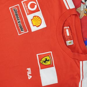 Könnte beinhalten: Rotes T-Shirt mit weißen Akzenten und Sponsorenlogos, darunter Vodafone, Shell und Bridgestone. Mit Ferrari-Logo und Fila-Branding. Das Shirt hat einen Rundhalsausschnitt und weiße Streifen an der Seite.