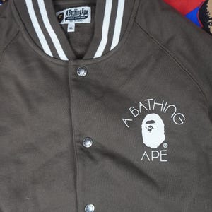 May include: A brown zip-up jacket with a white and brown striped collar. The jacket features the text "A Bathing Ape" and a graphic of an ape's face. The jacket has a button closure and is labeled XL.