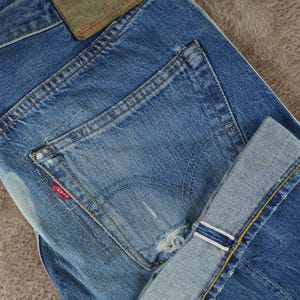 May include: Blue denim jeans featuring a distressed finish, a back pocket with a small red Levi's brand tag, a worn leather-like patch on the waistband, and a cuffed hem revealing a white and blue selvedge edge with yellow contrast stitching.