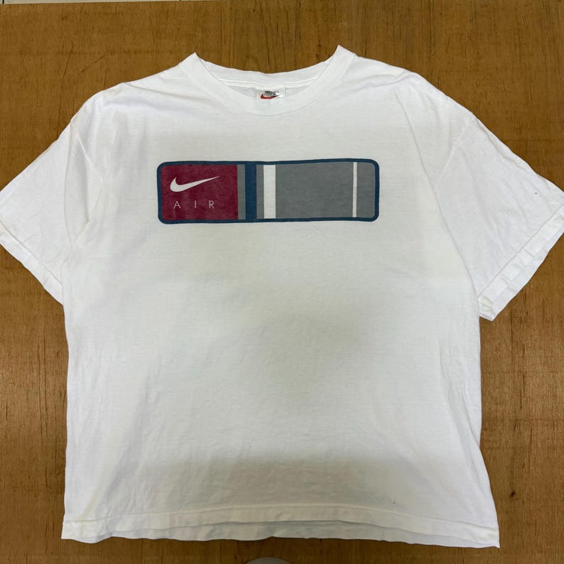 Op de afbeelding: Wit T-shirt met een rechthoekige afbeelding op de voorkant. De afbeelding heeft een rood, blauw en grijs ontwerp met het woord "AIR" en het Nike swoosh-logo. Het shirt heeft korte mouwen en een ronde hals.