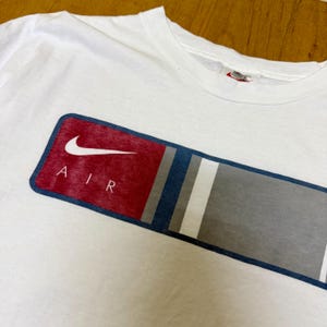 Op de afbeelding: Wit T-shirt met een rechthoekige afbeelding. De afbeelding heeft een rood en grijs gedeelte met het woord "AIR" en een witte swoosh-logo. Het ontwerp bevat ook blauwe en witte strepen. Klein Nike-logo bij de kraag.