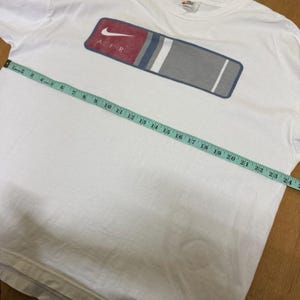 Op de afbeelding: Wit T-shirt met een rechthoekig ontwerp met de Nike swoosh en het woord "AIR". Het ontwerp bevat rode, blauwe, grijze en witte strepen. Een meetlint ligt over het shirt.