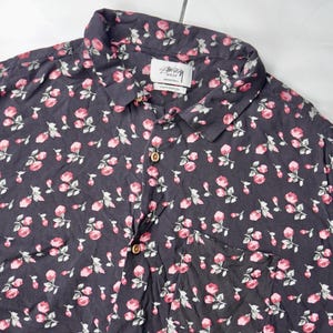 May include: A dark gray button-up shirt with a floral pattern of pink roses. The shirt has a collar and a button closure. The brand name "Stussy" is visible on the label.