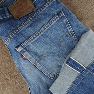 Puede incluir: Vaqueros de mezclilla azul con una etiqueta de cuero color canela con el texto Levi Strauss & Co. Original Clothing Every Garment Guaranteed W30 L36. Los vaqueros tienen un bolsillo trasero con costuras amarillas, una etiqueta roja y dobladillo.