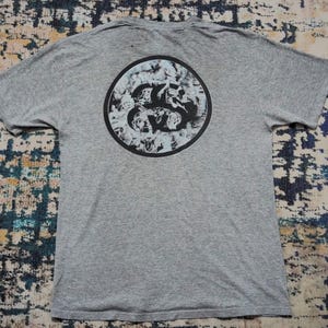 May include: A grey t-shirt featuring a circular graphic on the back. The graphic is a black circle with a white and black design. The shirt has short sleeves and a crew neck.