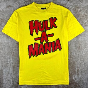 Op de afbeelding: Een felgeel T-shirt met korte mouwen en de tekst HULK-A-MANIA in dikke, grillige rode letters met een zwarte omlijning in het midden van de borst. Het T-shirt heeft een standaard ronde hals en een rechte zoom aan de onderkant.