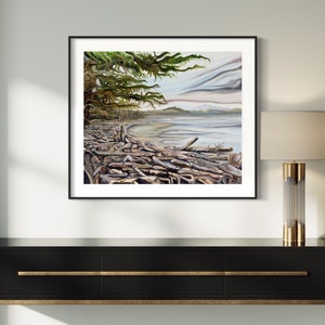 May include: A framed landscape painting depicts a coastal scene with driftwood, a sandy beach, and a body of water under a cloudy sky. The artwork features a large tree with green foliage on the left side. The frame is black.