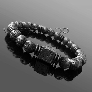 Könnte beinhalten: Schwarzes Lava-Stein-Armband mit einem quadratischen schwarzen Lava-Stein-Anhänger. Das Armband liegt auf einer reflektierenden Oberfläche.