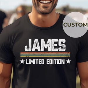 May include: Black t-shirt with the name "JAMES" in white, distressed lettering. Below the name are horizontal stripes in orange, yellow, green, and blue. The text "LIMITED EDITION" is below the stripes, with a star on each side.