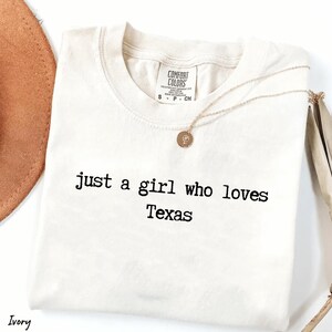 May include: Cream-coloured t-shirt with the text "just a girl who loves Texas" in black. A gold necklace with a pendant rests on the shirt. A brown hat is partially visible in the background.