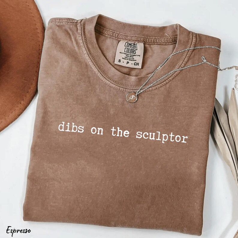 Op de afbeelding: Espresso-kleurig t-shirt met de witte tekst "dibs on the sculptor". Het shirt heeft een ronde hals en een klein label op de kraag. Een zilveren ketting met een hanger is over het shirt gedrapeerd.