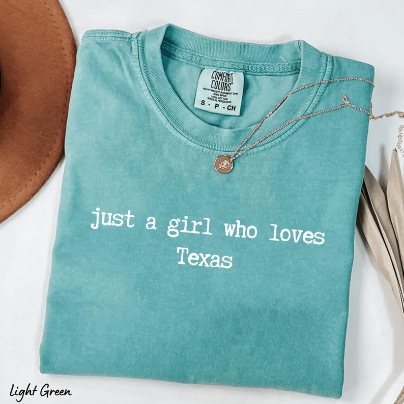 May include: Light green t-shirt with the text "just a girl who loves Texas" in white. The shirt has a crew neck and short sleeves. A gold necklace with a pendant is draped across the shirt. A brown hat is visible in the upper left corner.