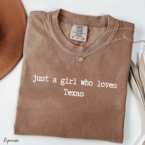 May include: Espresso-coloured t-shirt with the text "just a girl who loves Texas" in white. The shirt has a crew neck and short sleeves. A silver necklace with a pendant is draped across the shirt.