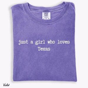 May include: A violet-coloured t-shirt featuring the white text "just a girl who loves Texas". The shirt has a crew neck and short sleeves. The brand label "Comfort Colors" is visible at the top.