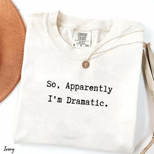 May include: Ivory-colored t-shirt with the text "So, Apparently I'm Dramatic." A gold-colored necklace with a pendant is draped across the shirt. The shirt is folded, and a portion of a brown hat is visible.