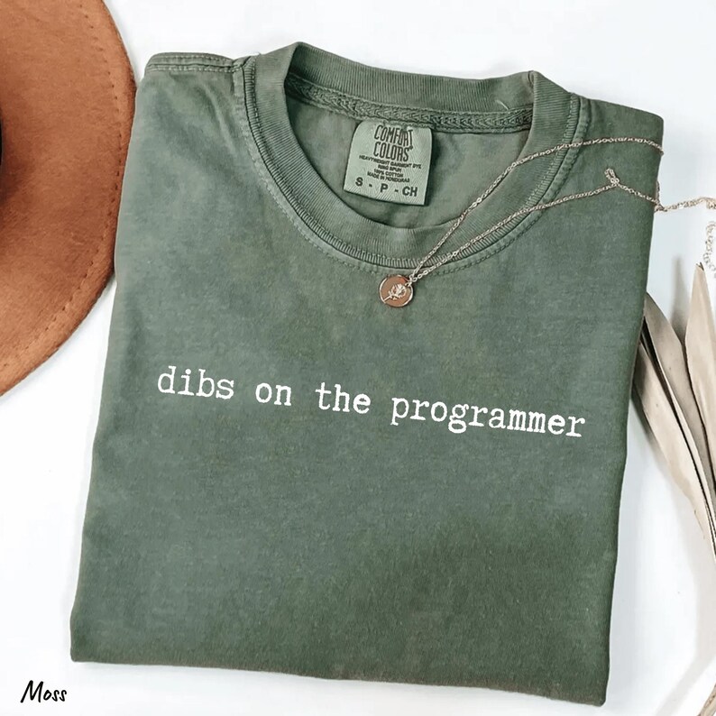 Pu&ograve; includere: Una maglietta verde muschio con la scritta bianca "dibs on the programmer". La maglietta &egrave; piegata e ha un'etichetta Comfort Colors. Una collana d'argento con un piccolo ciondolo &egrave; appoggiata sulla maglietta.