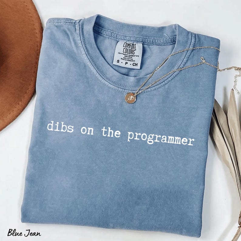Pu&ograve; includere: Una maglietta blu jeans con la scritta "dibs on the programmer" stampata in bianco. Una collana d'oro con un ciondolo &egrave; drappeggiata sulla maglietta. La maglietta &egrave; piegata e ha un'etichetta Comfort Colors.