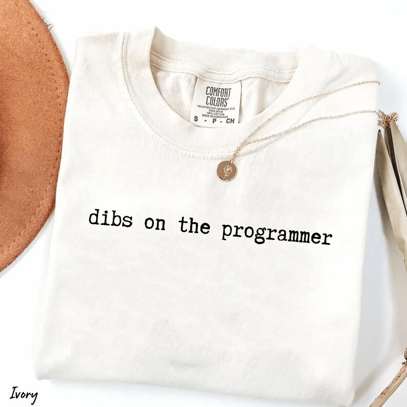 Pu&ograve; includere: T-shirt color avorio con la scritta nera "dibs on the programmer". Una collana d'oro con un ciondolo &egrave; appoggiata sulla maglietta. La maglietta ha un'etichetta Comfort Colors.