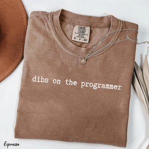 May include: Espresso-coloured t-shirt with the phrase "dibs on the programmer" in white. The shirt has a crew neck and short sleeves. A silver necklace with a pendant is draped across the shirt. The shirt is folded on a white surface.